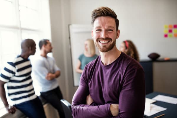 un homme au premier plan avec un grand sourire et 4 personnes en réunion brainstorming derrière